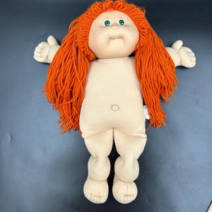 May include: A vintage doll with a soft, flesh-toned body and bright orange yarn hair. The doll has a round face with green eyes and a small, open mouth. Its arms and legs are outstretched, and it has a small belly button. The doll is a classic toy.