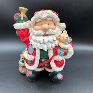 May include: A festive Santa Claus figurine, featuring a red suit with white trim, a long white beard, and a rosy nose. He holds a small bell and a teddy bear. The figurine has a patchwork design on the suit and black boots.