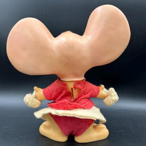 Op de afbeelding: Vintage speelgoedfiguur met grote, afgeronde oren en een rode vilten outfit. De figuur heeft een rode top, een rode short en een witte rok. De armen hebben witte pompons. De figuur is gemaakt van lichtbruin plastic.
