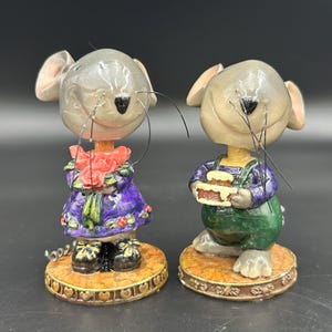 May include: Two whimsical mouse figurines. One mouse wears a purple dress and holds a bouquet of pink roses. The other mouse wears green overalls and holds a cake. Both stand on decorative circular bases.