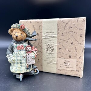 May include: A Lang & Wise collectible figurine of a teddy bear wearing a plaid dress and hat, holding a snowman. The figurine is on a tricycle and comes with its original box. The box has the text "Teddys & Toys" and "Fine Gifts".