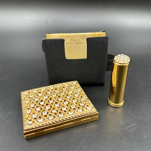 May include: A vintage gold-toned compact, lipstick, and a black case with the text "S&H FIFTH AVENUE". The compact is rectangular and covered in clear rhinestones. The lipstick and compact are gold-toned.