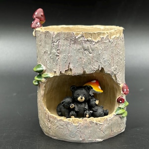 May include: A decorative tree stump-shaped planter with a family of black bears inside. The planter has a textured, gray-brown exterior with red and white mushrooms and green foliage accents. The bears are nestled under a mushroom.