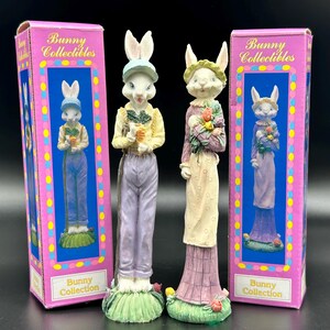 May include: Two bunny figurines, one holding carrots and the other flowers, stand next to their boxes. The boxes are pink with blue and yellow accents and the words "Bunny Collectibles" and "Bunny Collection". The bunnies are wearing hats and outfits.