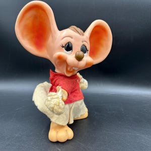 Op de afbeelding: Een vintage muisbeeldje met grote oren, gekleed in een rood shirt, beige shorts en een bijpassende hoed. De muis heeft een vrolijke uitdrukking en houdt een klein voorwerp in zijn handen. Het beeldje is gemaakt van een lichtgekleurd materiaal.