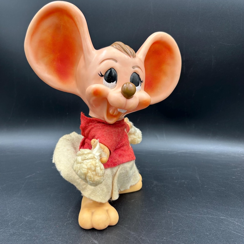 Op de afbeelding: Een vintage muisbeeldje met grote oren, gekleed in een rood shirt, beige shorts en een bijpassende hoed. De muis heeft een vrolijke uitdrukking en houdt een klein voorwerp in zijn handen. Het beeldje is gemaakt van een lichtgekleurd materiaal.