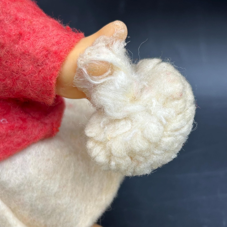 Op de afbeelding: Close-up van een poppenhand die een pluizig wit object vasthoudt. De arm van de pop is bedekt met een rood viltmateriaal. De hand van de pop is gemaakt van een glad, lichtgekleurd materiaal.