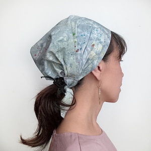 May include: A grey bandana with colourful splatter paint accents tied around a person's head. The bandana is tied in a knot at the back of the head and the ends are tucked under the knot.