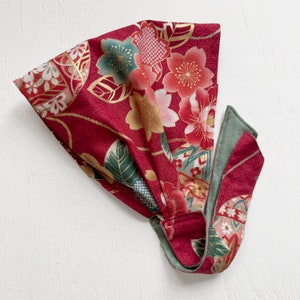 May include: A red fabric headband with a floral pattern in shades of pink, green, and gold. The headband has a long tie that can be tied in a bow.