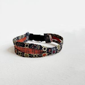 May include: A fabric headband with a black and floral pattern, accented with red and tan details. The headband has a black adjustable clasp. The floral pattern includes red, white, and yellow flowers.