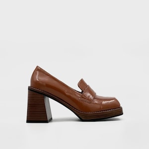 Könnte beinhalten: Brauner Leder-Loafer mit eckiger Spitze und klobigem Absatz in Holzoptik. Der Schuh hat ein glänzendes Finish und ein klassisches Penny-Loafer-Design. Der Absatz misst ca. 7,6 cm.