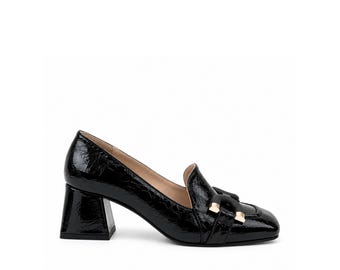 Könnte beinhalten: Ein schwarzer Loafer aus Lackleder mit knittriger Textur, ausgestattet mit einer quadratischen Schuhspitze, einem Blockabsatz und einem dekorativen schwarzen Riemen über dem Vorderblatt, akzentuiert durch zwei goldfarbene Metallelemente.