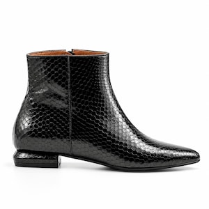 Könnte beinhalten: Eine schwarze Stiefelette mit glänzender Oberfläche und einer strukturierten Wabenmusterung. Der Schuh verfügt über eine spitz zulaufende Kappe, einen seitlichen Reißverschluss und einen flachen, rechteckigen Blockabsatz in passendem Schwarz.