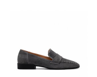 May include: A grey suede loafer featuring a low black heel and a pointed toe. The shoe is accented with prominent white whipstitching along the vamp and the top edge of the tongue, providing a distinct contrast against the dark grey material.