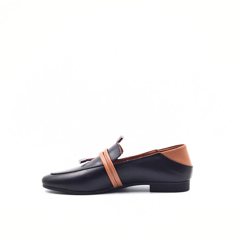 May include: Black leather loafer with a tan leather accent on the heel and a tan strap across the top. The shoe has a small silver tassel and a low heel. The shoe is a classic style.