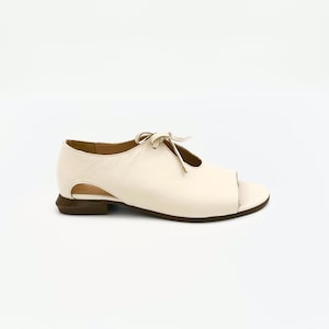 May include: Cream-coloured open-toe shoe with a small heel. The shoe features a cut-out design on the side and a bow detail on the top. The sole is a dark brown colour, and the shoe appears to be made of leather.