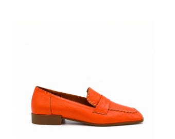 May include: A bright orange leather loafer featuring a square toe, a low brown block heel, and decorative whipstitch detailing along the vamp and apron. The shoe has a smooth finish and a structured silhouette with a classic penny loafer strap.