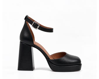 May include: A black platform shoe with a square toe, a high block heel, and an adjustable ankle strap featuring a silver-toned metal buckle. The interior lining of the shoe is a warm brown color, contrasting with the smooth black exterior.