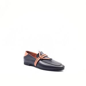May include: Black leather loafer with a square toe, tan leather heel detail, and tan leather tie accent. The shoe has a low heel and a minimalist design, suitable for casual or semi-formal wear.