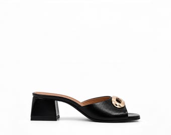 May include: A black mule sandal featuring a block heel and an open toe design. The upper strap is adorned with a large, polished gold-tone metallic chain link ornament. The interior footbed is a tan color, providing a contrast to the black exterior.