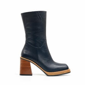 Könnte beinhalten: Ein wadenhoher Stiefel aus glattem, dunkelblauem Leder mit quadratischer Zehenpartie und einem dicken Blockabsatz aus hellem Holz. Der Stiefel verfügt über eine markante Holzplateausohle mit sichtbarer Maserung, die einen Kontrast zum dunklen Obermaterial bildet.