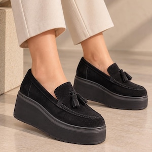 Op de afbeelding: Een paar zwarte suède loafers met een dikke plateauzool en decoratieve kwastjes op de wreef. De schoenen hebben een mocassin-stijl stiksel op de neus en een effen zwarte afwerking, wat zorgt voor een opvallend en robuust silhouet.
