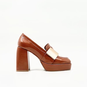 Può includere: Mocassino marrone con tacco a plateau, punta quadrata e dettaglio dorato. La scarpa ha un tacco spesso e una finitura lucida. La suola platform aggiunge altezza. Uno stile classico con un tocco moderno.