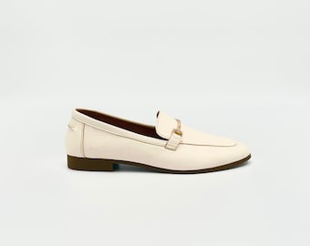 Puede incluir: Un par de mocasines de cuero de color blanco roto con un detalle de hebilla dorada. Los mocasines tienen una suela marrón.