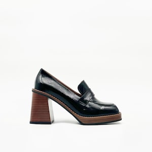 May include: Black leather heeled loafer with a square toe and a chunky, brown wooden heel. The shoe has a classic penny loafer design with a glossy finish and a wooden sole.