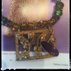 May include: A vintage necklace featuring a gold-toned pendant with floral details and a large, oval purple stone. The necklace has a beaded design with gold, green, and purple beads. The background includes text and a patterned design.