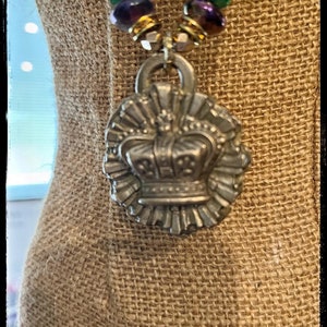 May include: A silver-toned pendant necklace featuring a detailed crown design. The pendant is suspended from a beaded strand with green, purple, and black beads, accented by gold-colored spacers. The necklace is displayed on a burlap-textured surface.