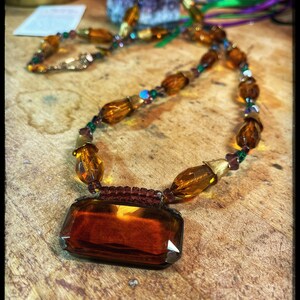 May include: A vintage necklace featuring a large, rectangular amber-colored pendant. The necklace has a beaded design with amber-colored, oval-shaped beads and smaller, colorful accent beads. The necklace is displayed on a wooden surface.