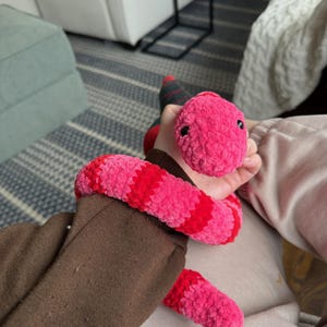 Könnte beinhalten: Ein rosa und rotes gehäkeltes Schlangenspielzeug mit schwarzen Augen. Die Schlange liegt auf dem Schoß einer Person.