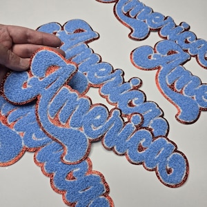 May include: Blue felt patches spelling "America" in a script font. Each patch has a red glitter border and a white outline. The patches are scattered on a white surface.