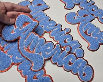 Peut inclure: Plusieurs écussons en feutre bleu avec le mot "America" écrit en script. Les écussons ont une bordure rouge pailletée et un contour blanc. Ils sont posés sur une surface blanche.