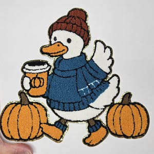 May include: A whimsical patch featuring a cartoon duck in a blue sweater, brown hat, and boots, holding a pumpkin spice latte. Two orange pumpkins are at the duck's feet. The patch has a gold glitter outline.