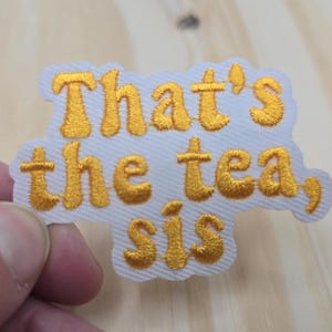 May include: Embroidered patch with the phrase "That's the tea, sis" in gold thread on a white background. The patch has a scalloped edge and is held by a hand.