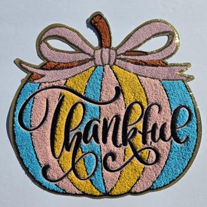 May include: A colorful pumpkin-shaped decoration with the word "Thankful" in elegant script. The pumpkin features vertical stripes in pink, yellow, and blue, topped with a pink bow and a brown stem. The edges are outlined in gold.