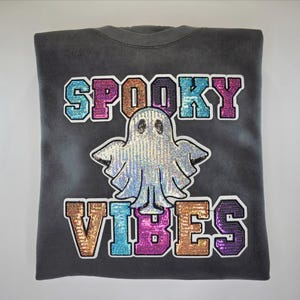 May include: Dark grey sweatshirt featuring the words "SPOOKY VIBES" in colourful sequined letters. A sequined ghost graphic is centered below the word "SPOOKY". The sweatshirt is folded and displayed on a white surface.