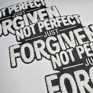 May include: Black and white patches with the text "NOT PERFECT JUST FORGIVEN". The patches have a textured, fuzzy white font on a black background. The patches are angled on a white surface.