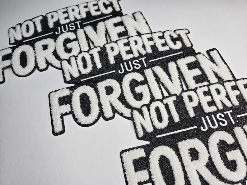May include: Black and white patches with the text "NOT PERFECT JUST FORGIVEN". The patches have a textured, fuzzy white font on a black background. The patches are angled on a white surface.