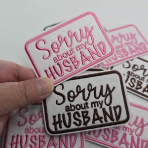May include: Embroidered patches with the phrase "Sorry about my husband" in various color combinations. The patches are rectangular with rounded corners and feature white fabric with pink or brown lettering and borders. A hand holds a patch.