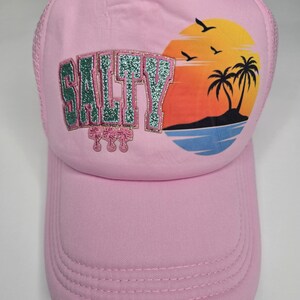 May include: Pink trucker hat with the word "SALTY" in green glitter lettering. The hat features a sunset graphic with palm trees, birds, and ocean waves. The hat has a curved brim and mesh back.
