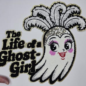 Peut inclure: Écusson brodé représentant un fantôme de dessin animé avec un couvre-chef à paillettes et des cheveux en plumes. Le fantôme a des joues roses, de longs cils et un sourire rouge. Le texte "The Life of a Ghost Girl" est brodé en noir avec une bordure dorée.
