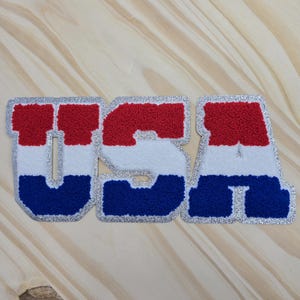 May include: A USA patch in red, white, and blue, with silver glitter edging. The letters are displayed on a light wood grain background. This patch is designed for application to clothing or accessories.