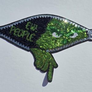 May include: A sequined patch in the shape of a zipper, featuring a green creature with white eyes and a pointing finger. The patch has the words "Ew, People" in green and silver lettering on a black background.