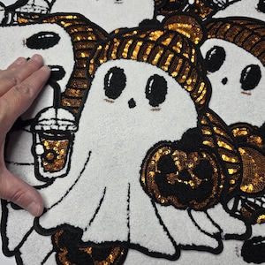 May include: White ghost-shaped patches with black eyes, holding a pumpkin and a drink. The ghost wears a gold sequined beanie. The patches have a black border and are decorated with gold sequins. Halloween-themed.
