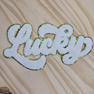 May include: White, fluffy "Lucky" patch with a green glitter border. The word is in a cursive font. The patch is on a light brown wooden surface.