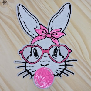 May include: A white bunny face patch with black outlines, wearing pink heart-shaped glasses and a pink bow. The bunny is blowing a pink bubblegum bubble. The patch is on a light wood surface.