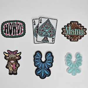 May include: A collection of embroidered patches with various designs. Includes a patch with the word "HOWDY", two playing cards with spade designs, a patch with the word "Mama", a cartoon cow, and two floral-shaped patches.